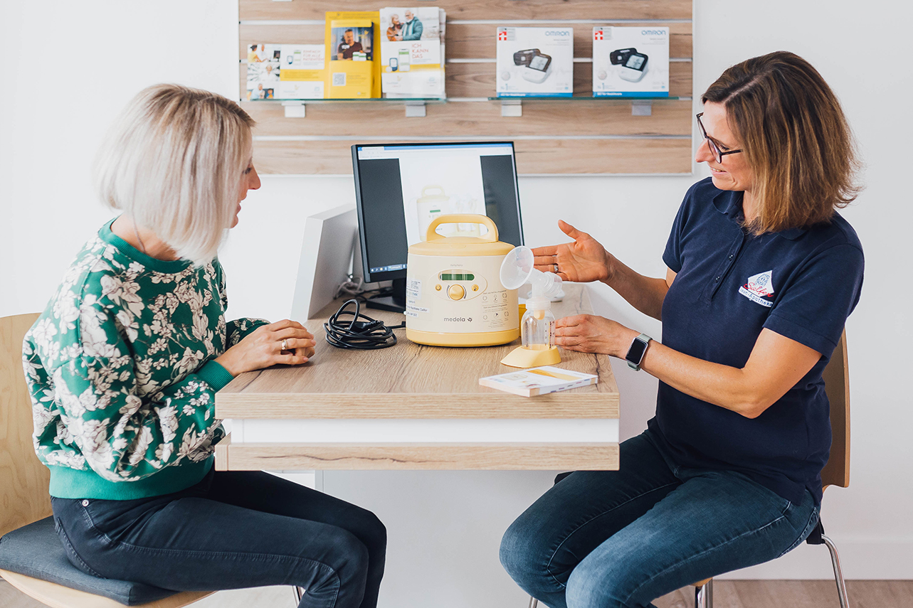 Mitarbeiterin sitzt mit einer Kundin am Tisch und zeigt ihr die Funktionen einer Milchpumpe