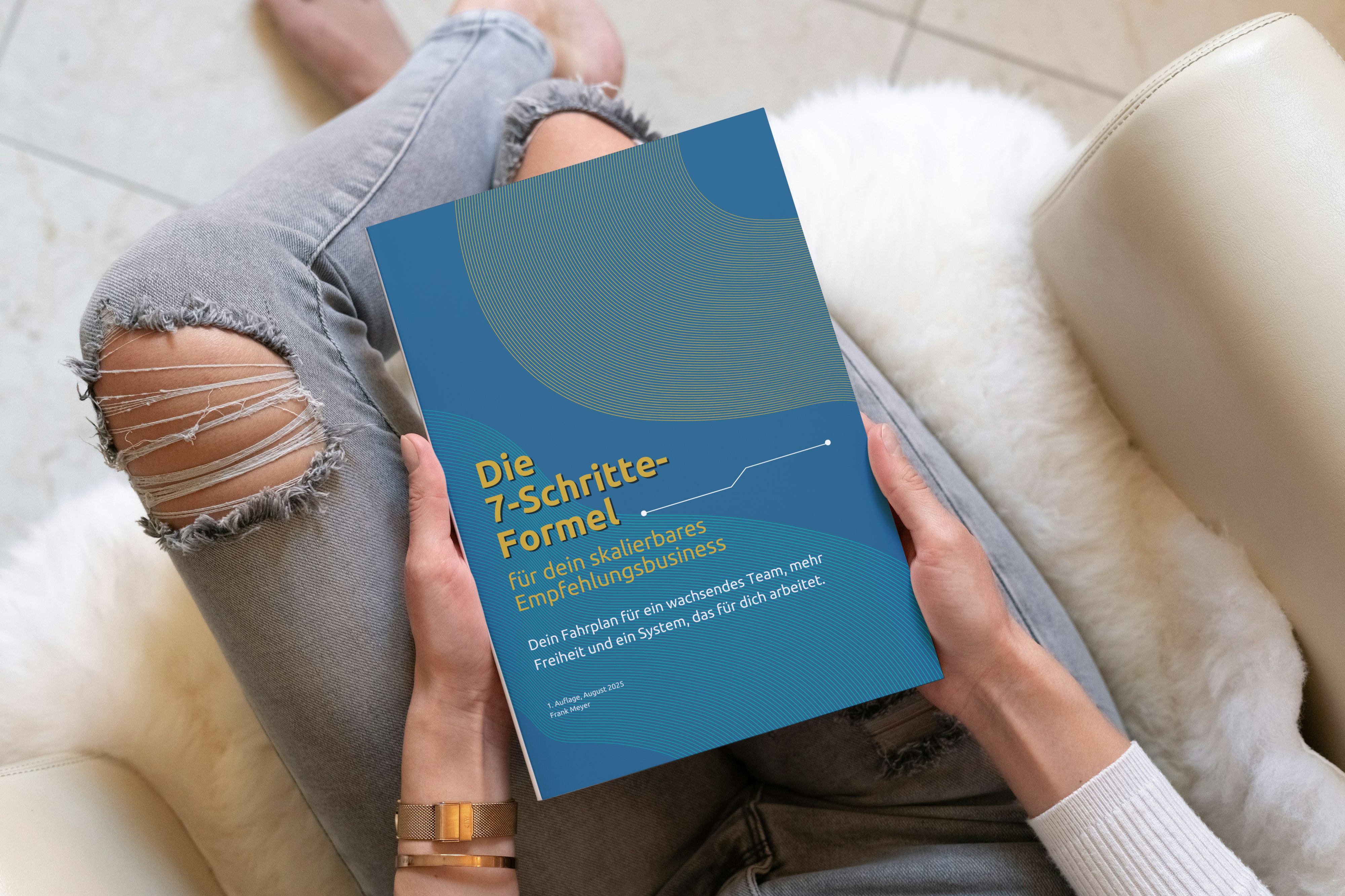 Person in grauer Jeans mit Rissen sitzt entspannt auf einem Sessel und hält den PDF-Report „Die 7-Schritte-Formel für dein skalierbares Empfehlungsbusiness“ in den Händen – Fahrplan für Teamaufbau, mehr Freiheit und ein automatisiertes System.