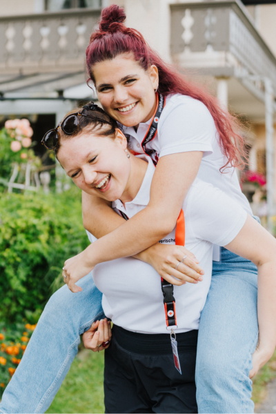 Zwei Fundraiserinnen bei ihrem Ferienjob im Freien, lächelnd und fröhlich. Eine trägt die andere huckepack. Beide tragen weiße Shirts und Schlüsselbänder. Der Hintergrund ist grün und blumig.