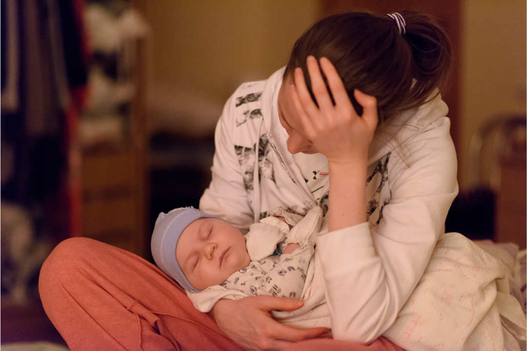 Eine Frau stützt dien Kopf in der Hand ab, sie wirkt erschöpft. Sie hält ein schlafendes Baby im Arm.