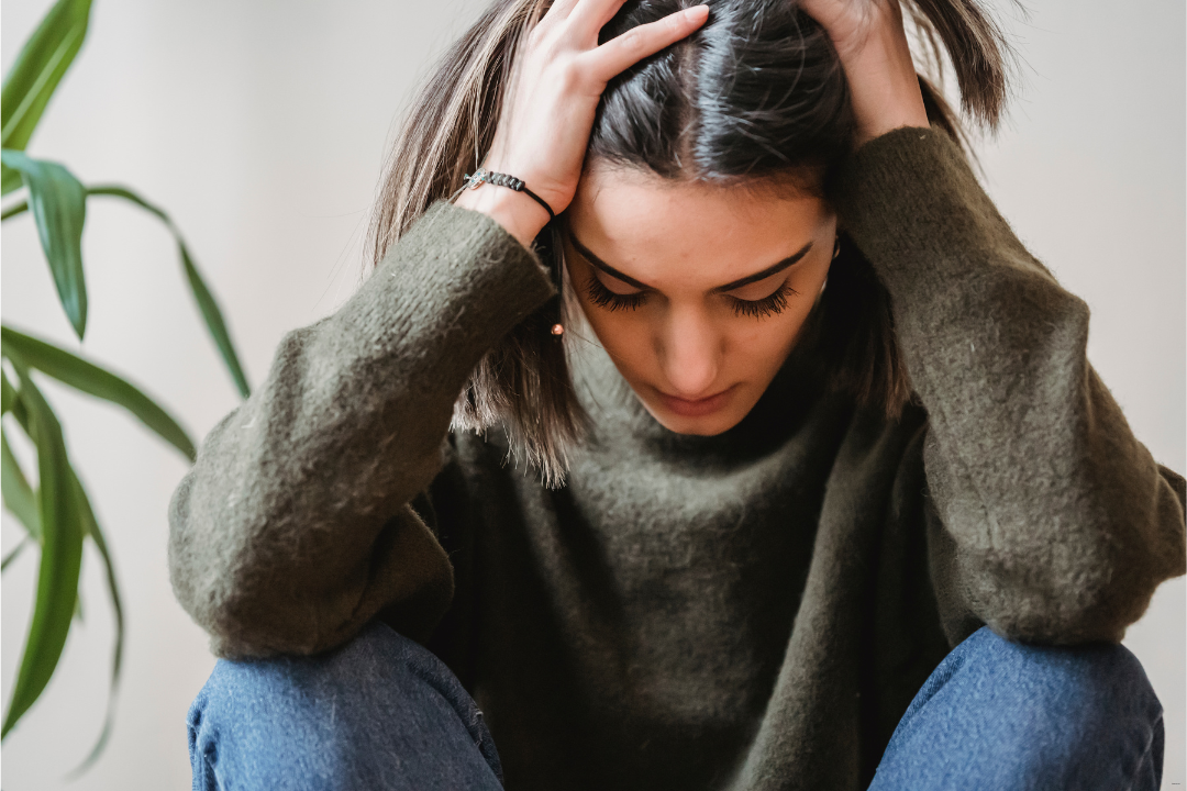 Eine Frau mit langen braunen Haaren stützt ihren Kopf in ihren händen ab. Sie wirkt müde, erschöpft. Sie trägt einen grünen Pullover und eine Jeans.. 