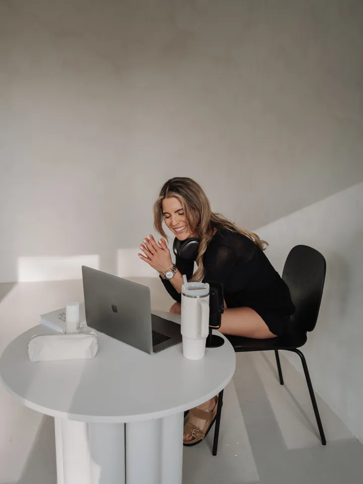Nina sitzt lachend am Laptop, vor sich ein Mikrofon, die Hände zusammengelegt.