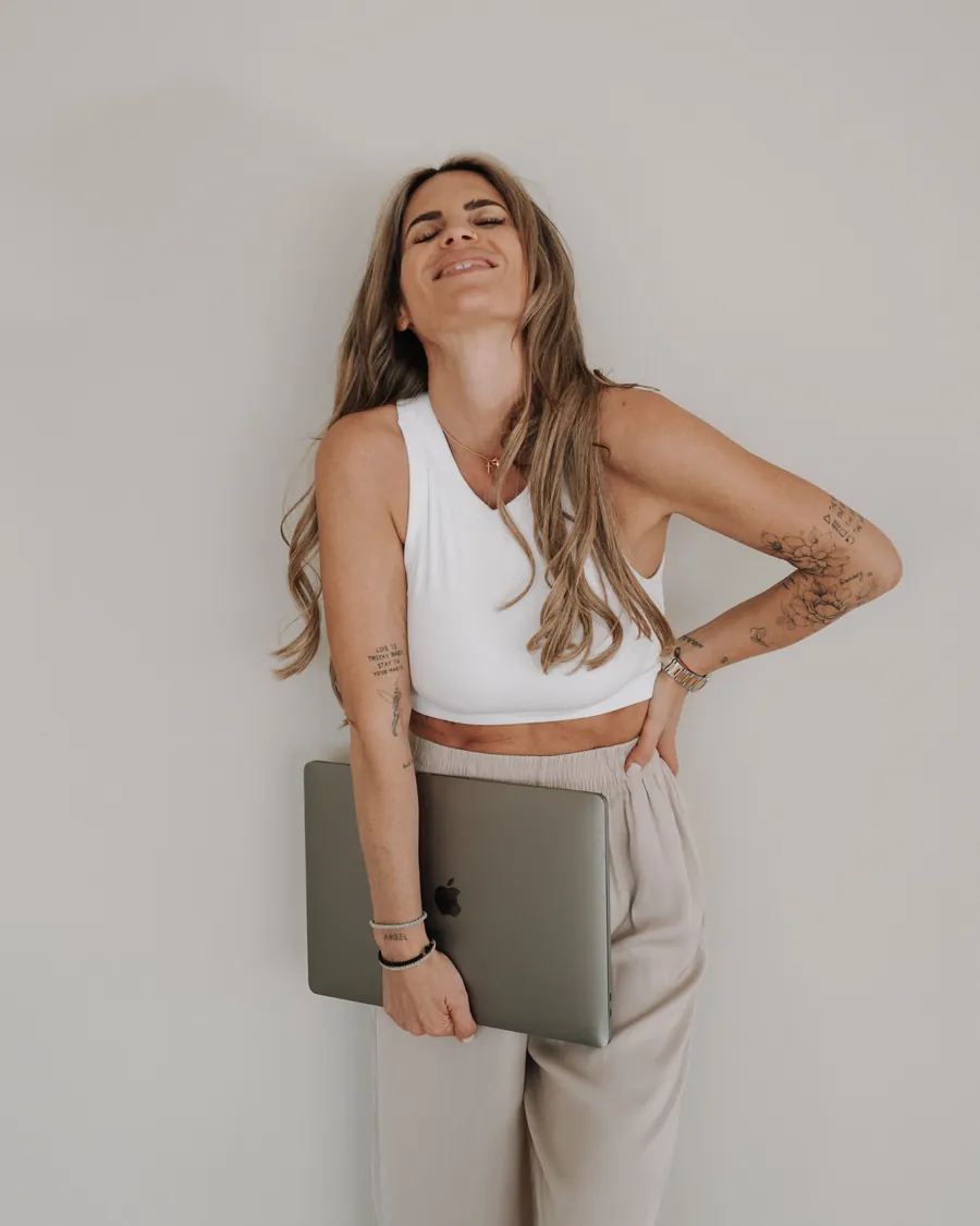 Nina lehnt lachend an einer hellen Wand, einen Laptop unter dem Arm.
