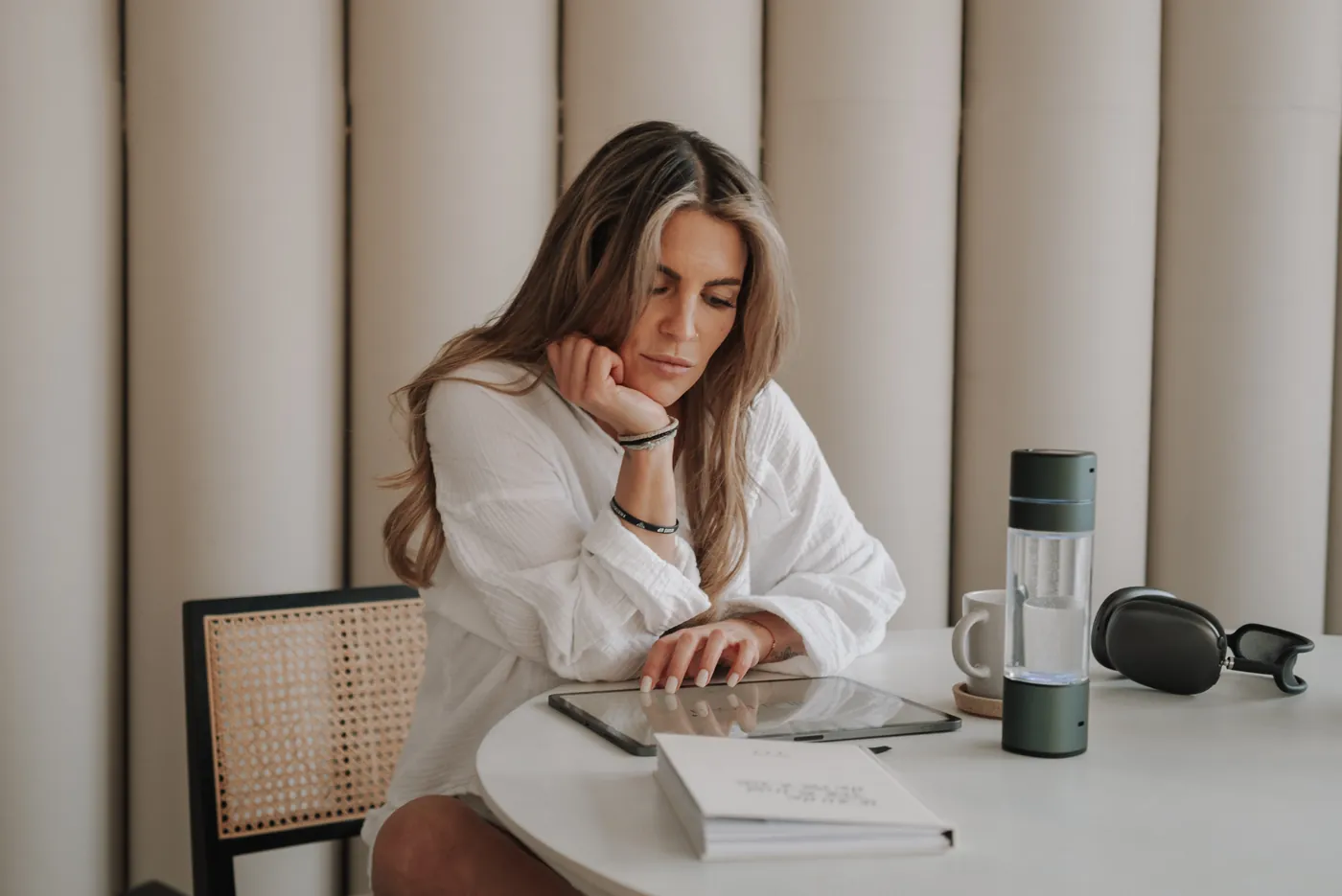 Nina in weißer Bluse sitzt nachdenklich an einem hellen Tisch, vor ihr Tablet und Notizbuch.