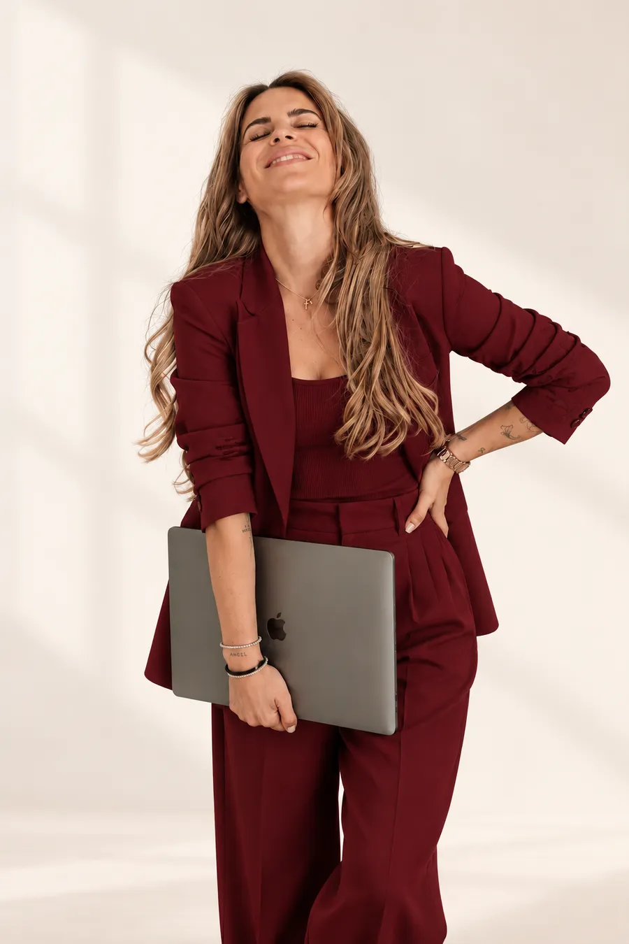 Nina im bordeauxroten Anzug mit Laptop unter dem Arm, lachend vor hellem Hintergrund.