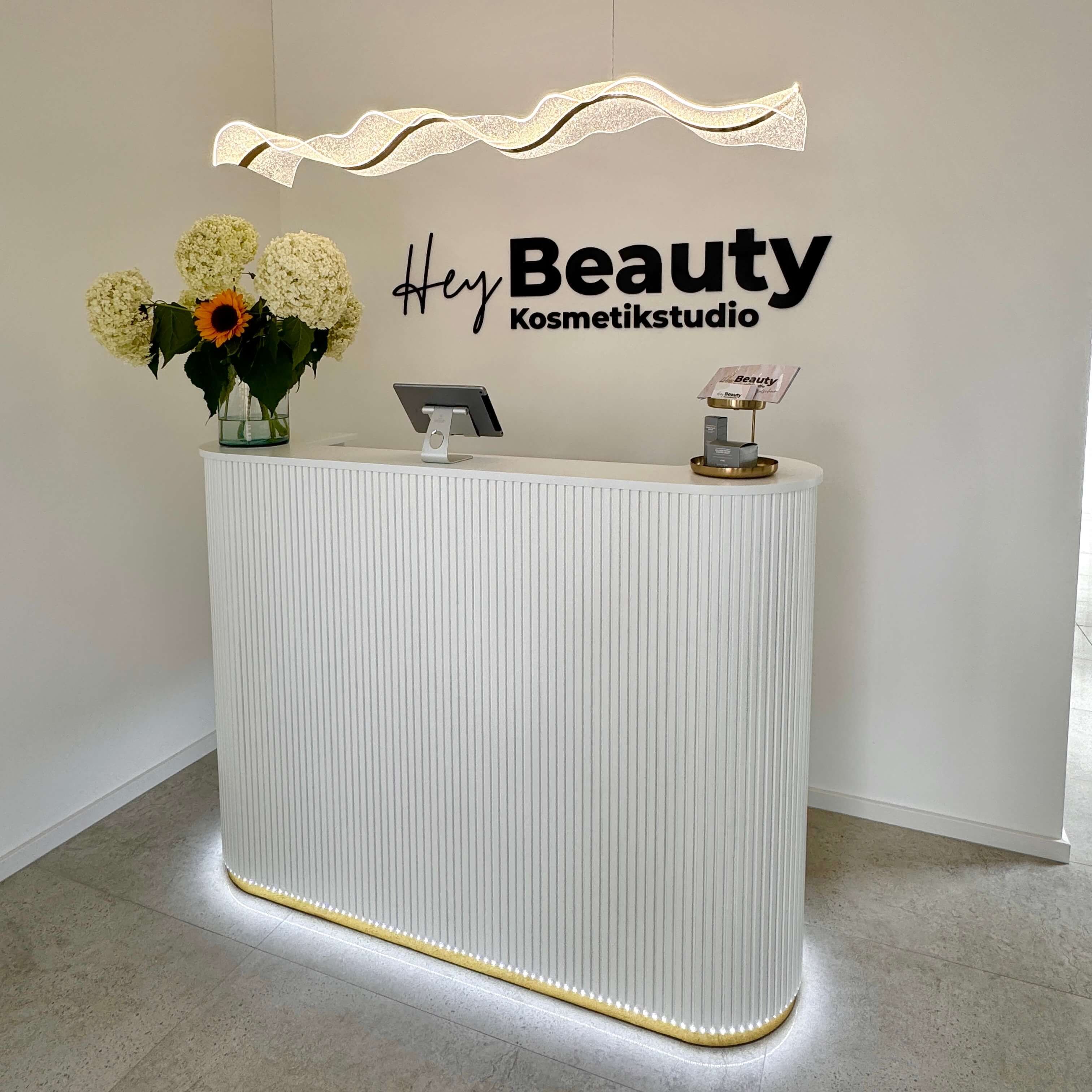 Empfangstheke mit beleuchtetem Logo von HeyBeauty Kosmetikstudio – minimalistisches Design in Weiß und Gold