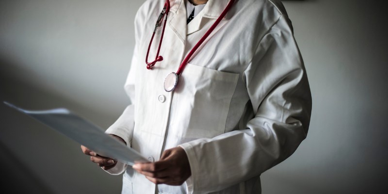 stehender Arzt mit weißem Kittel und Stethoskop, betrachtet in den Händen haltend ein Dokument