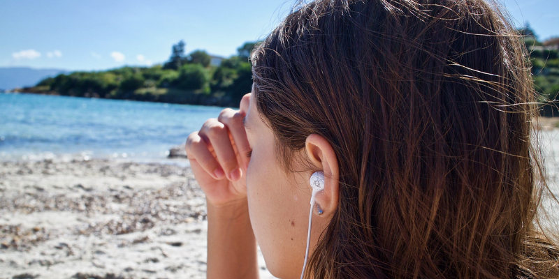 Frau mit Kopfhörer im Ohr sitzt am Strand, ihre rechte Hand liegt an der Stirn an