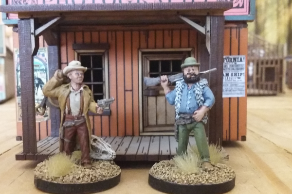 Zwei handbemalte Cowboy-Miniaturen vor einem Western-Saloon mit Waffen und Outfits