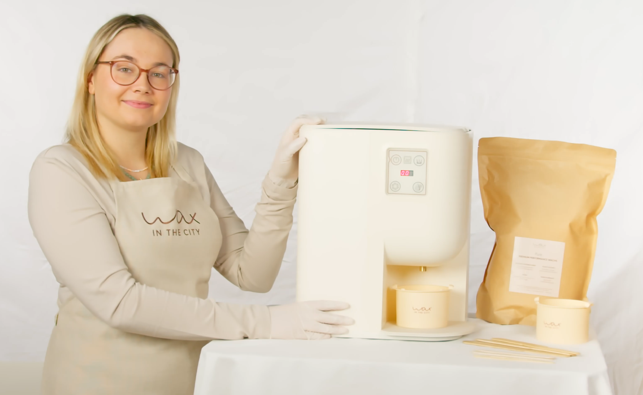 Maureen, Schulungsleiterin von Wax in the City, steht neben der WaxInside Waxing Station, die auf einem Tisch steht. Daneben befindet sich ein Beutel des PURE Wachs sowie zwei Silikon-Cups mit Spateln.